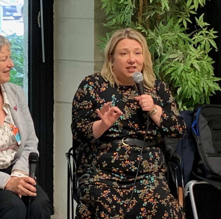 Gail Honeyman is sitting indoors and talking animatedly into a microphone to an audience. A woman is sitting next to her with a microphone in her lap, listening to Gail and smiling.